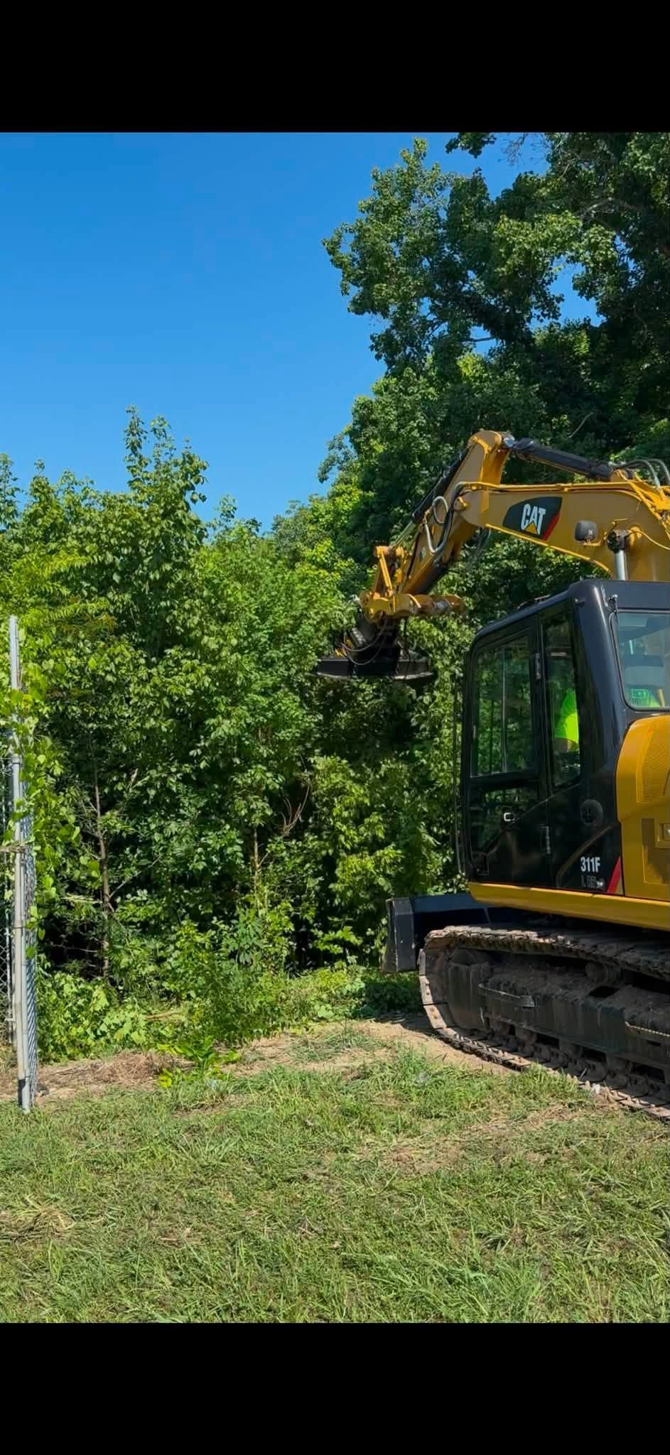 Yellow excavator clearing brush beside a dense green hedge under a bright blue sky