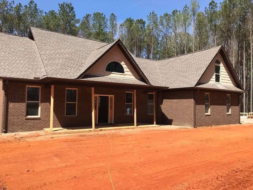 Brick house with a covered porch and gray roof on a red dirt lot, surrounded by trees