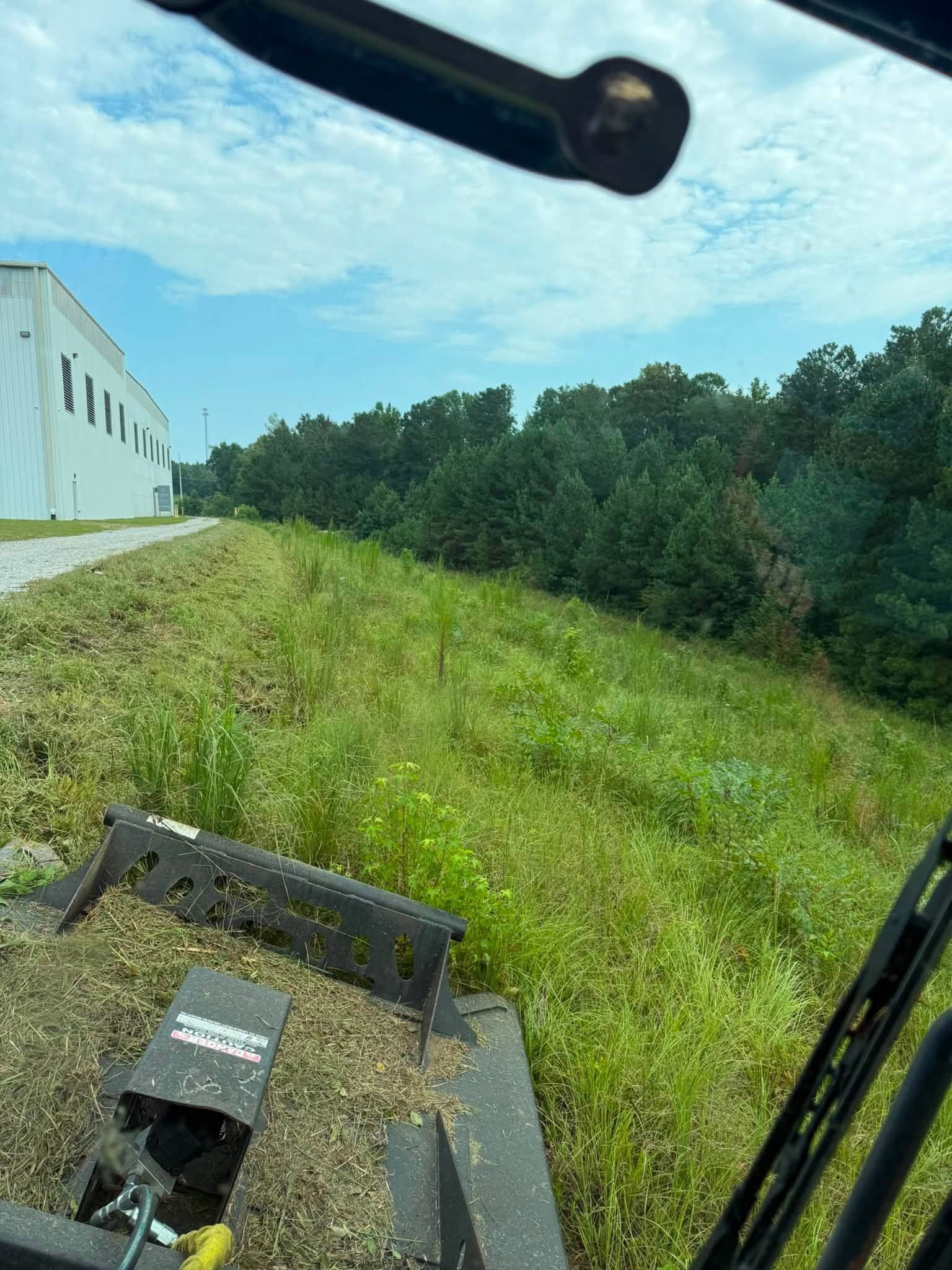 Grassy hillside beside a white building, viewed from a vehicle with a mower attachment in the foreground.