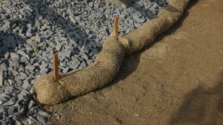 A coiled rope or log border separates gray gravel from tan sand, with two wooden stakes protruding.