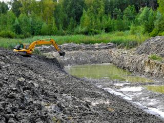 Excavator digging muddy earth beside a water-filled excavation pit, with trees in the background