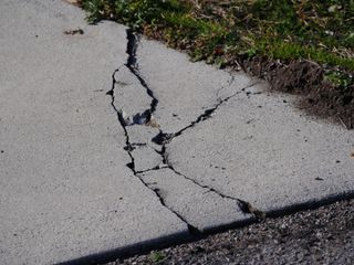 Cracked concrete sidewalk with large fissures and damaged pavement near grass edge