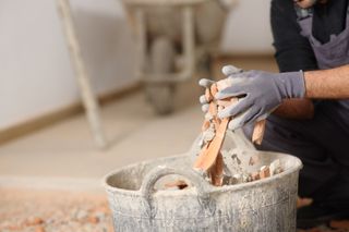 Gloved hands knead wet clay in a pottery bucket beside a worktable.