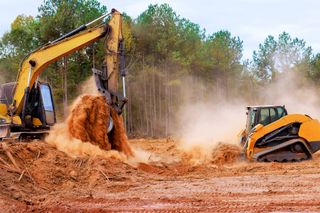 Excavator and bulldozer moving dirt at a dusty construction site with trees in the background