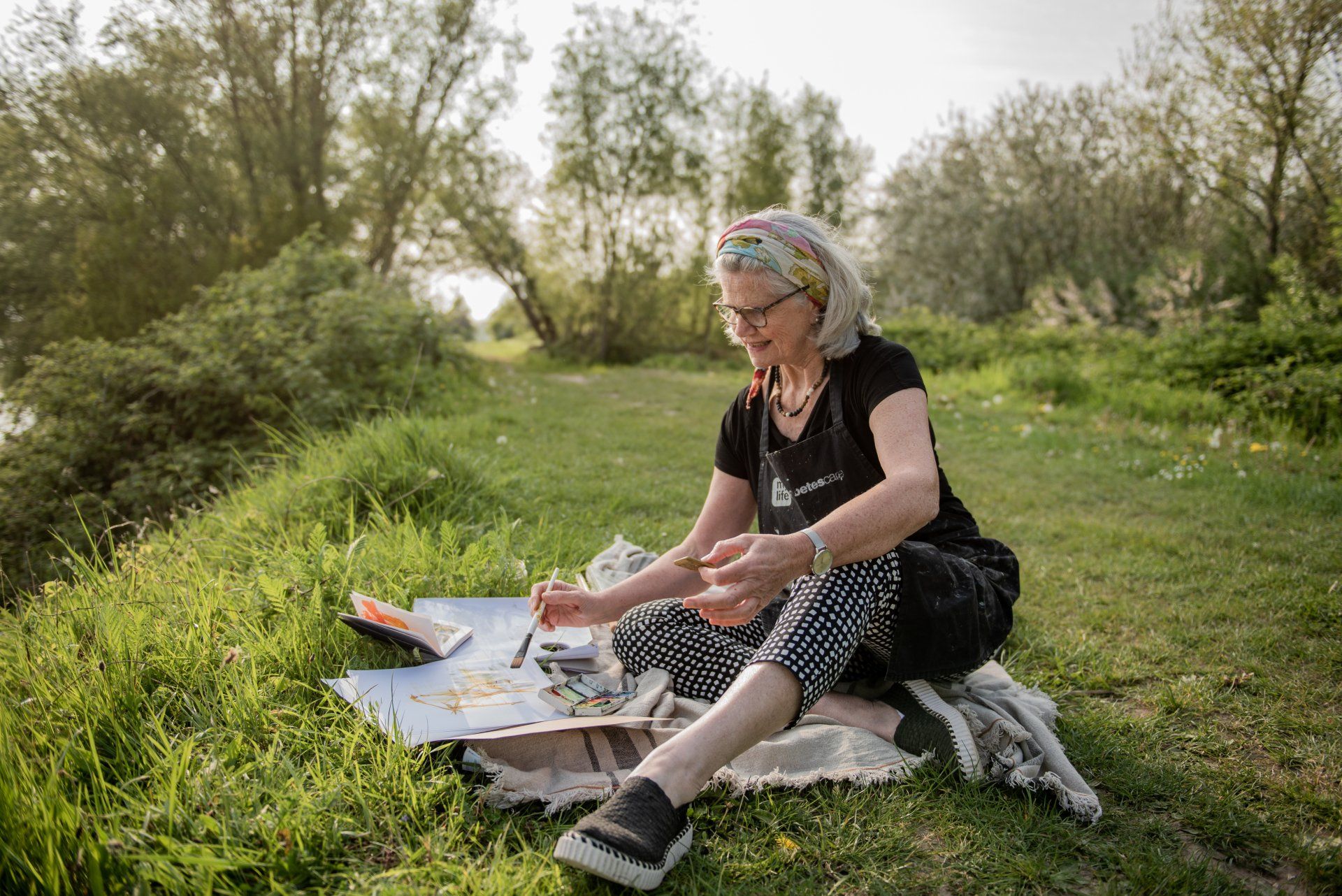 Vrouw schildert in de buitenlucht, zittend op een deken in een grasveldje vlakbij het water; ze draagt een hoofdband, een zwart shirt en een broek met een patroon.