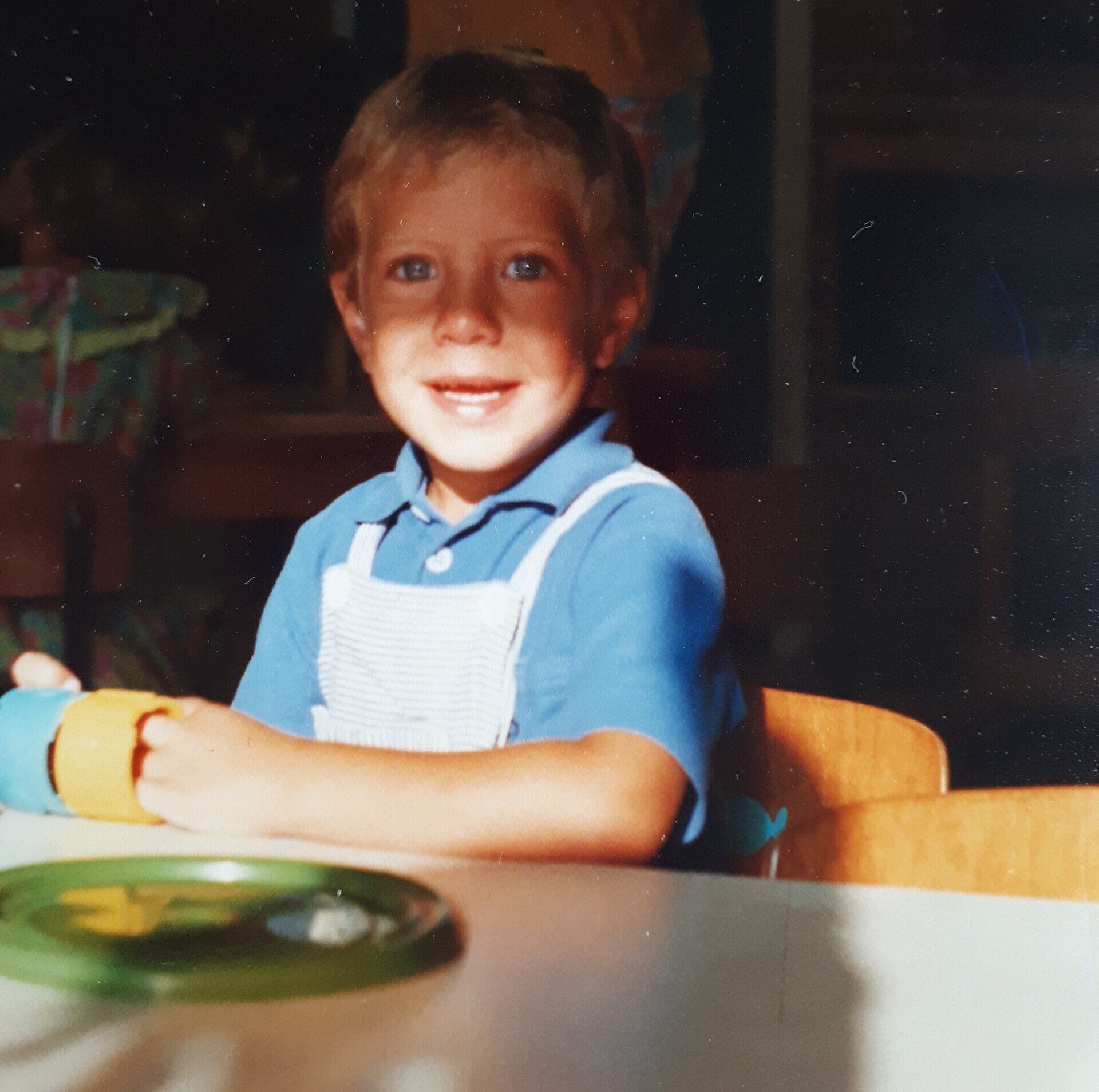Een lachend kind in een blauw shirt en een witte overall zit aan een tafel.