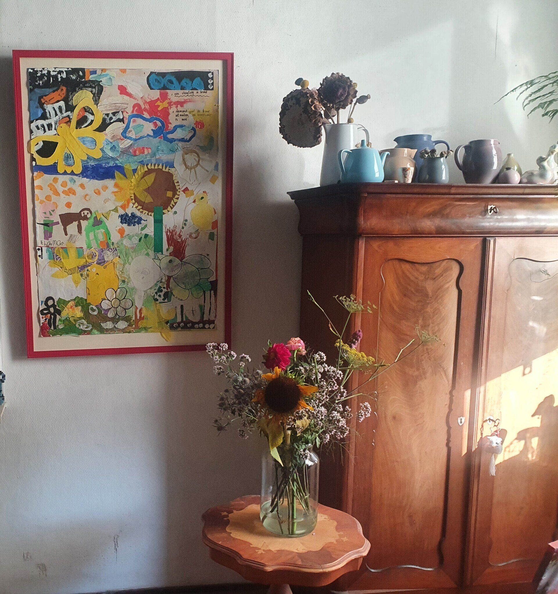 Aan een witte muur hangt een levendig schilderij naast een houten kast die is versierd met vazen ​​en bloemen.