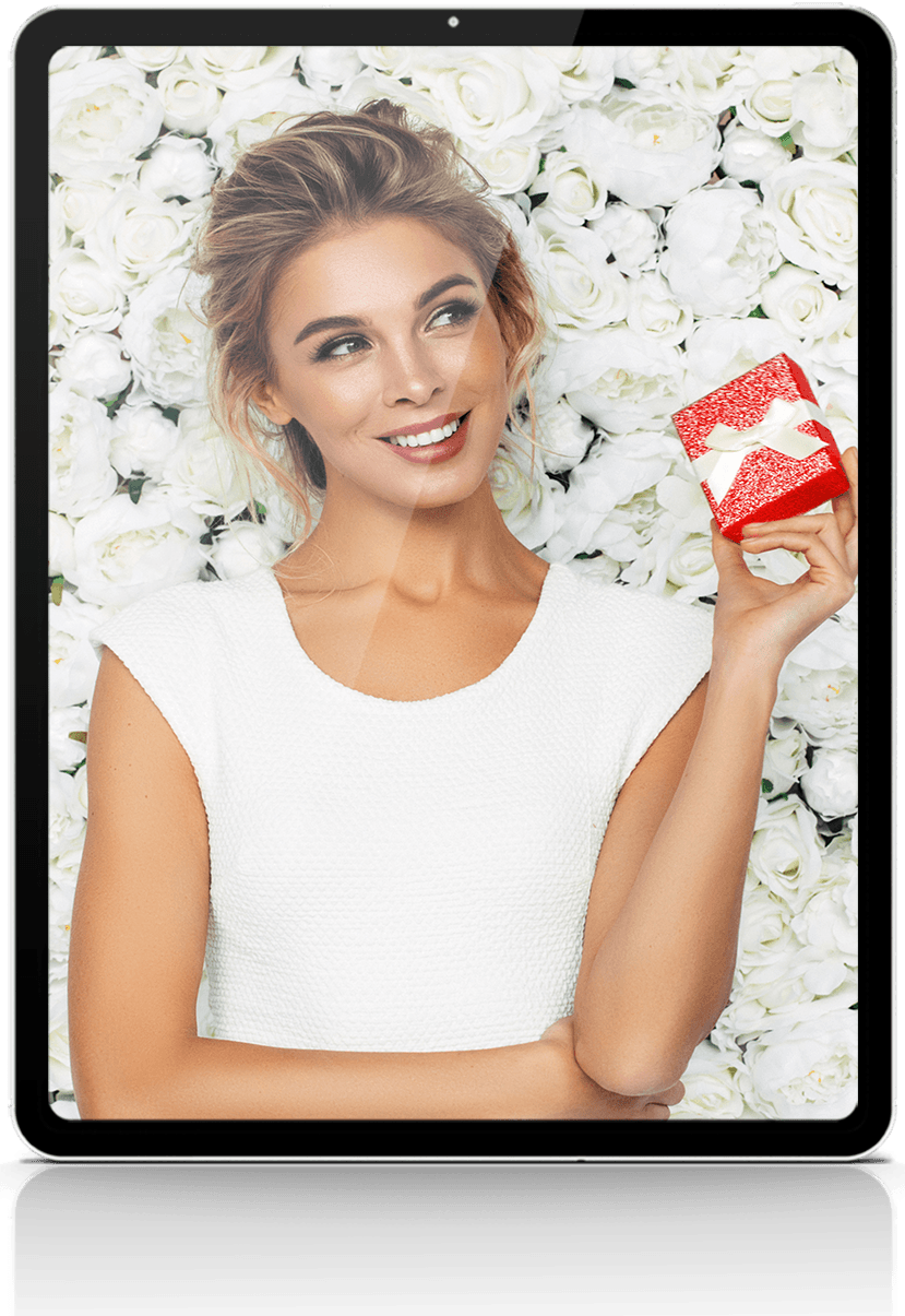 Woman in white dress smiles, holding red gift box, in front of white flower wall.