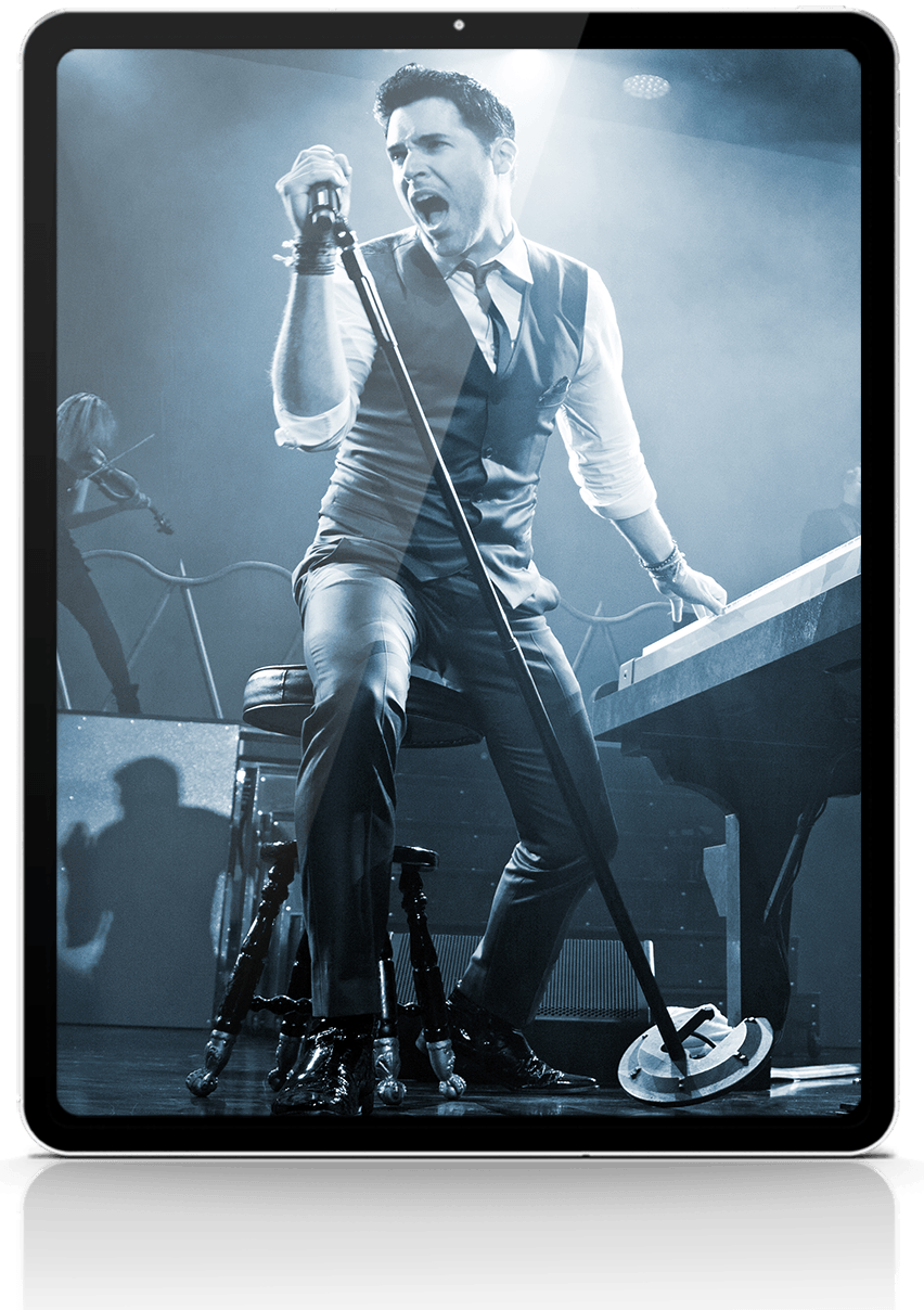 Man singing into microphone at a keyboard, stage setting.
