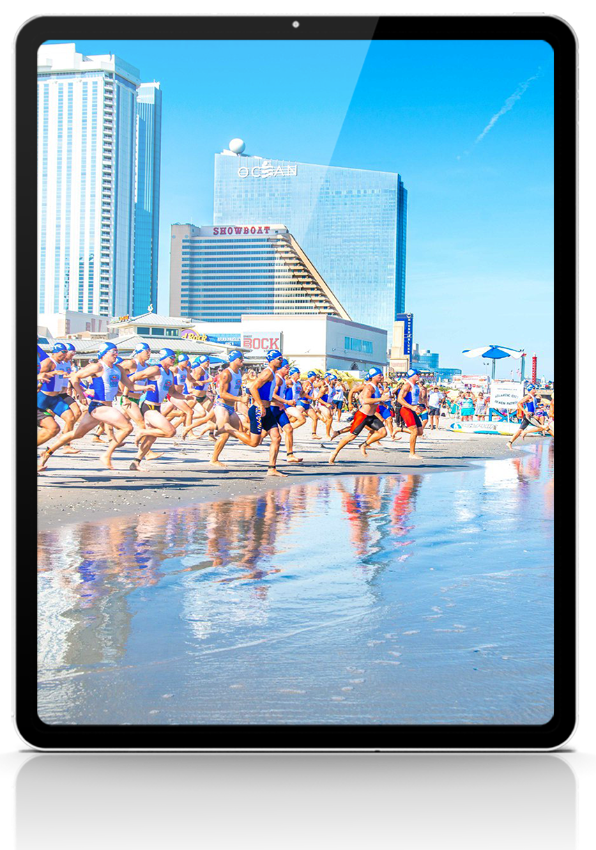People running on a beach in front of tall buildings, blue sky.