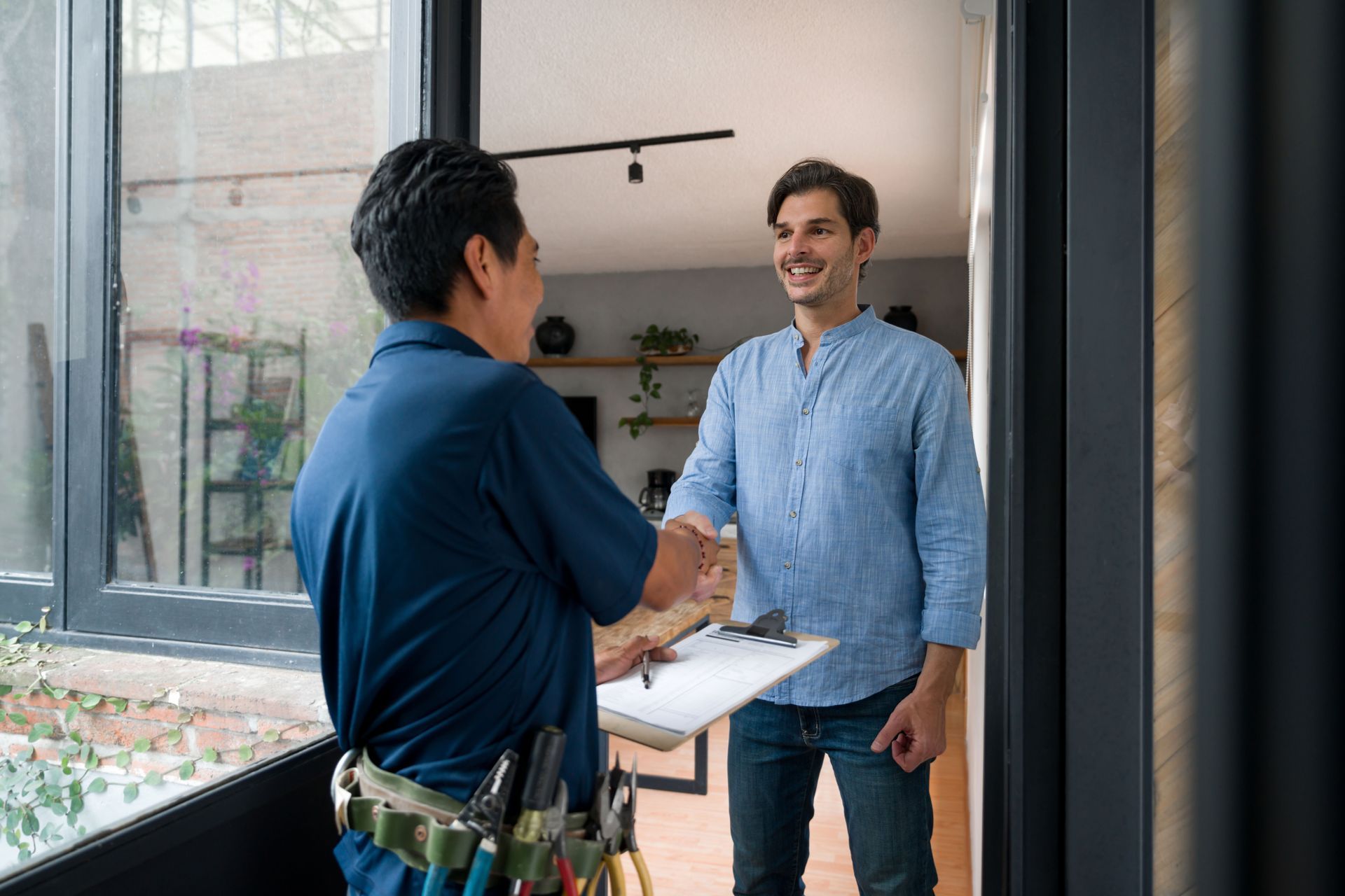 Two men shake hands at a doorway, one holding a clipboard in a bright office or shop entrance.