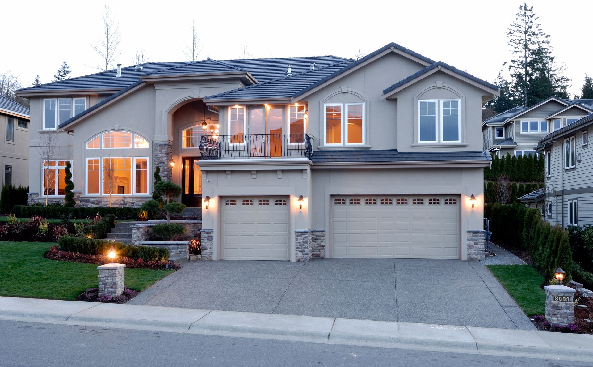 Large two-story suburban house with lit windows, double garage, and manicured front landscaping at dusk