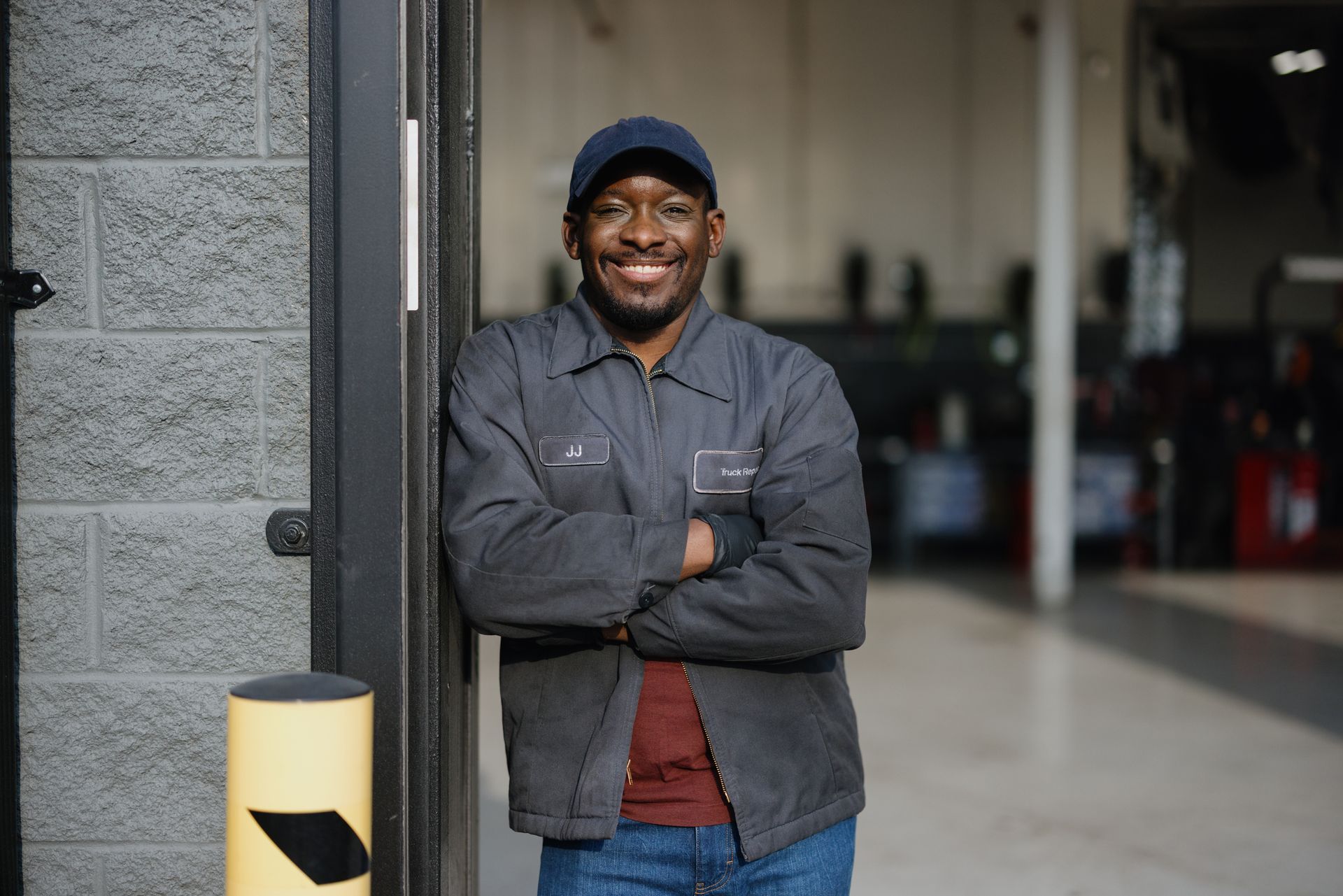 Man smiling with crossed arms in a workshop doorway, wearing a cap and dark jacket.
