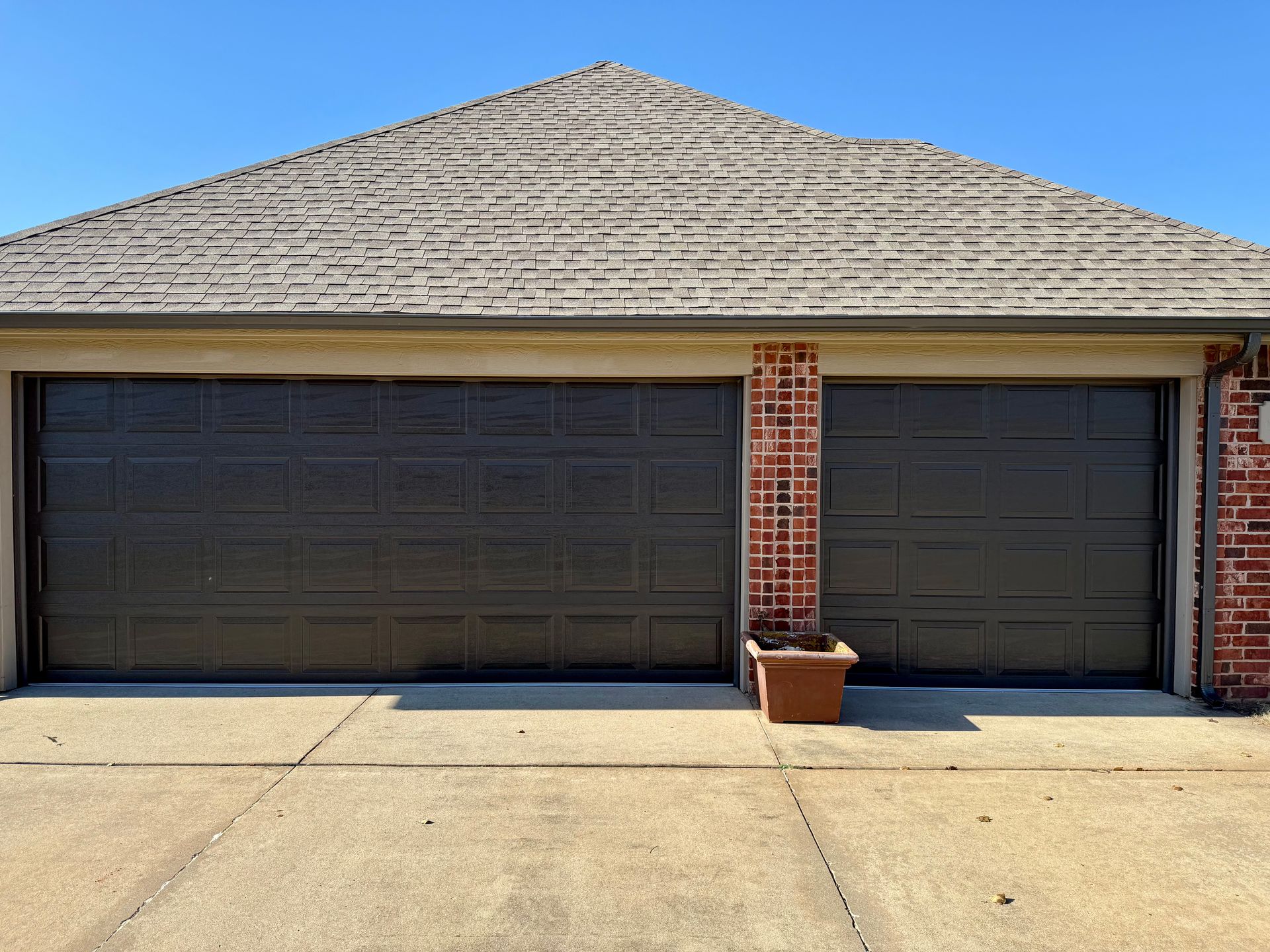 Two-car garage with dark doors, brick columns, and a tan shingle roof under a clear blue sky