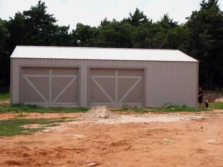 Gray metal two-bay garage/shed with white roof on a dirt lot, trees in the background