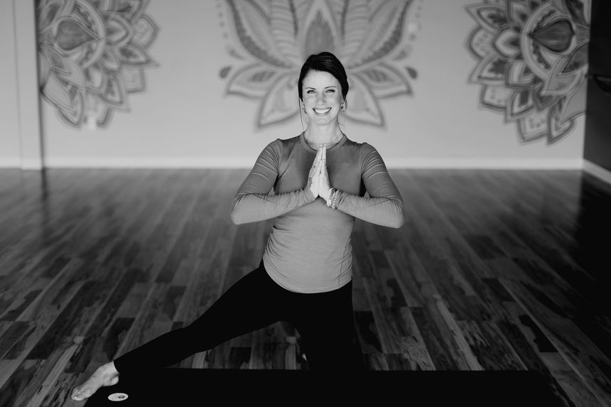 Woman in yoga pose, hands in prayer position, smiling, kneeling on mat in studio with mandala art.