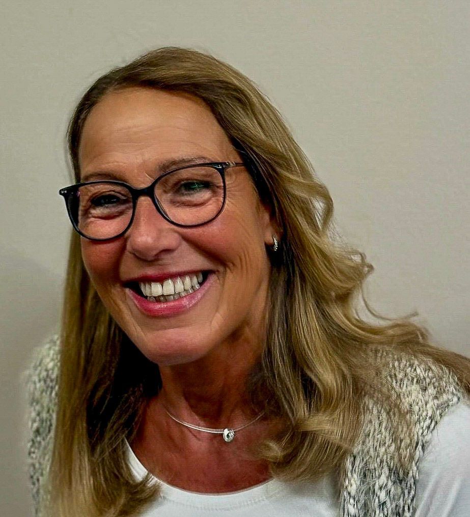Frau mit blonden Haaren, Brille, lächelnd, trägt ein weißes Oberteil und eine weiße Weste.