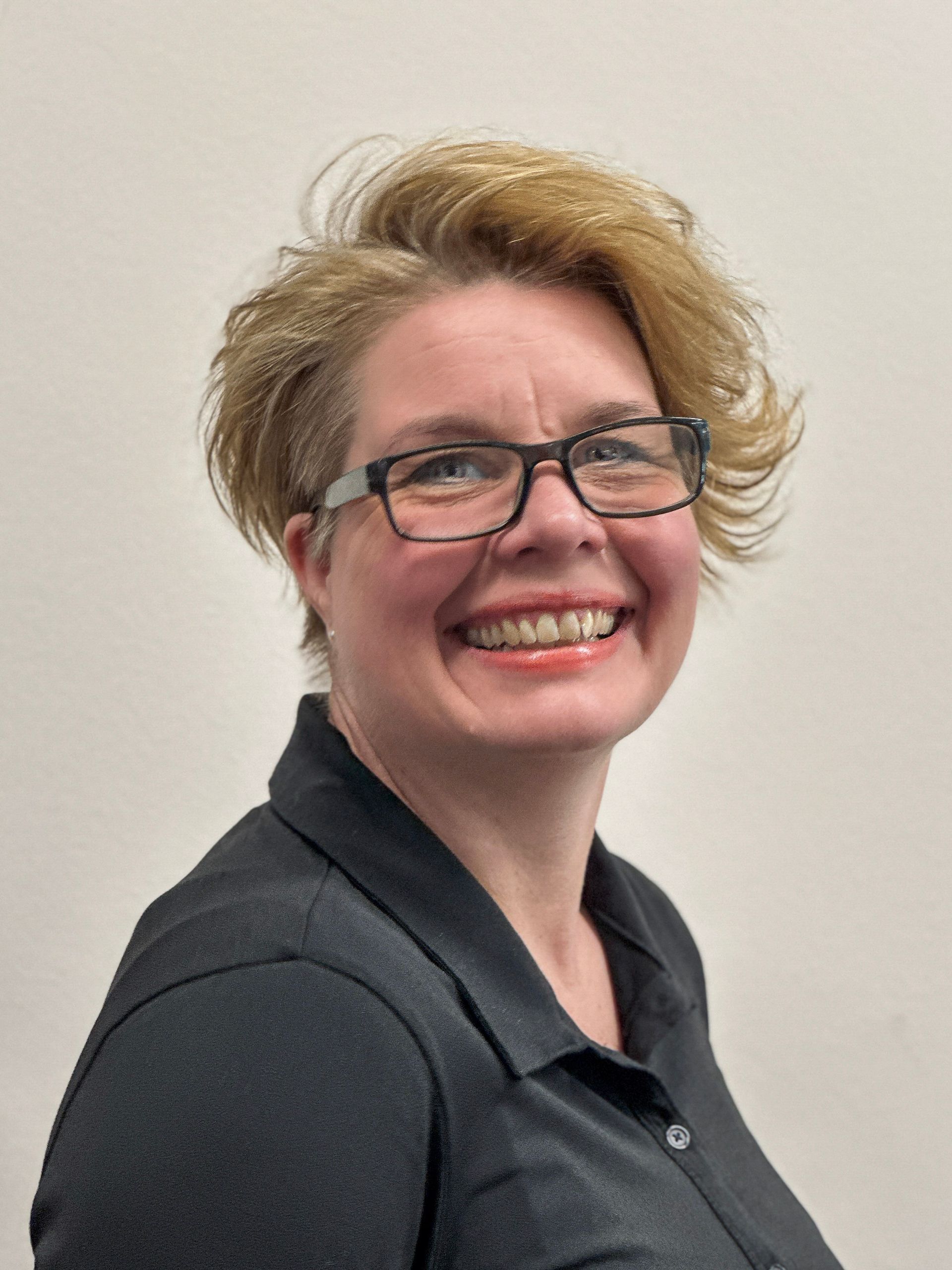 Frau mit blonden, kurzen Haaren, trägt eine Brille und lächelt. Dunkles Hemd, neutraler Hintergrund.