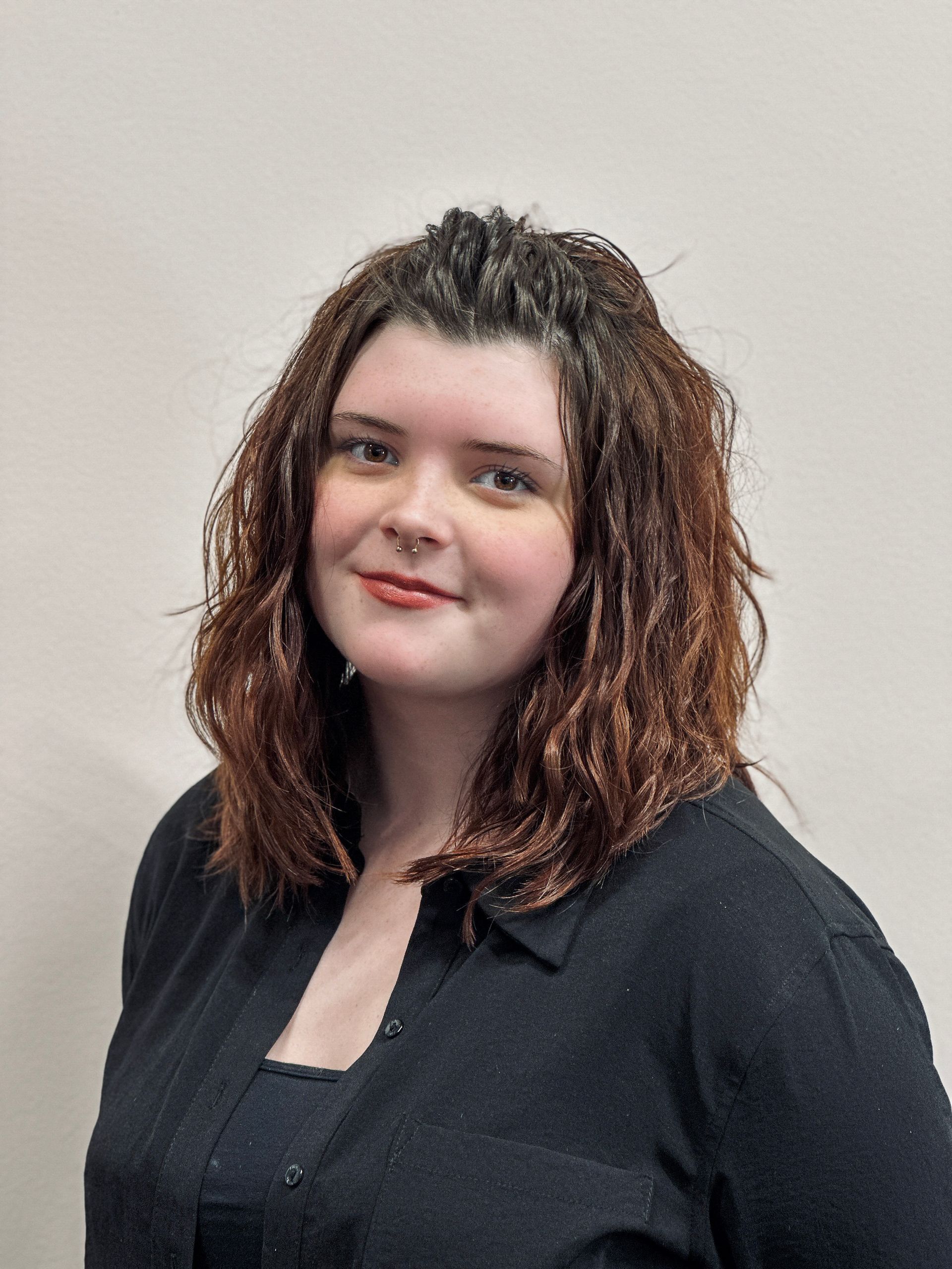 Frau mit braunen lockigen Haaren, lächelnd, trägt ein schwarzes Oberteil, vor einer hellen Wand.
