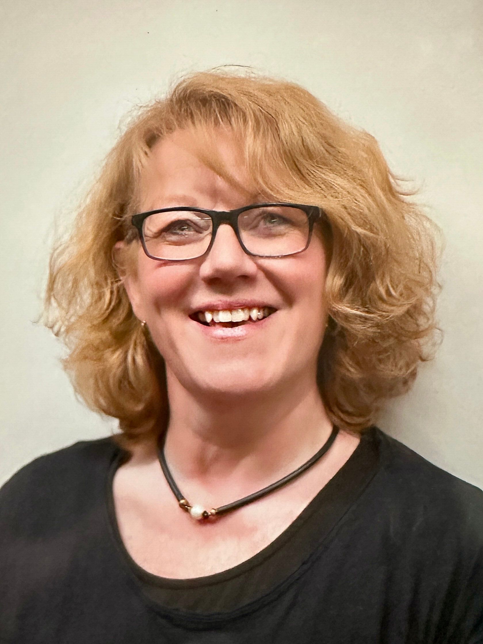 Frau mit Brille, blonden Locken und schwarzem Hemd lächelt in die Kamera. Schwarze Perlenkette.