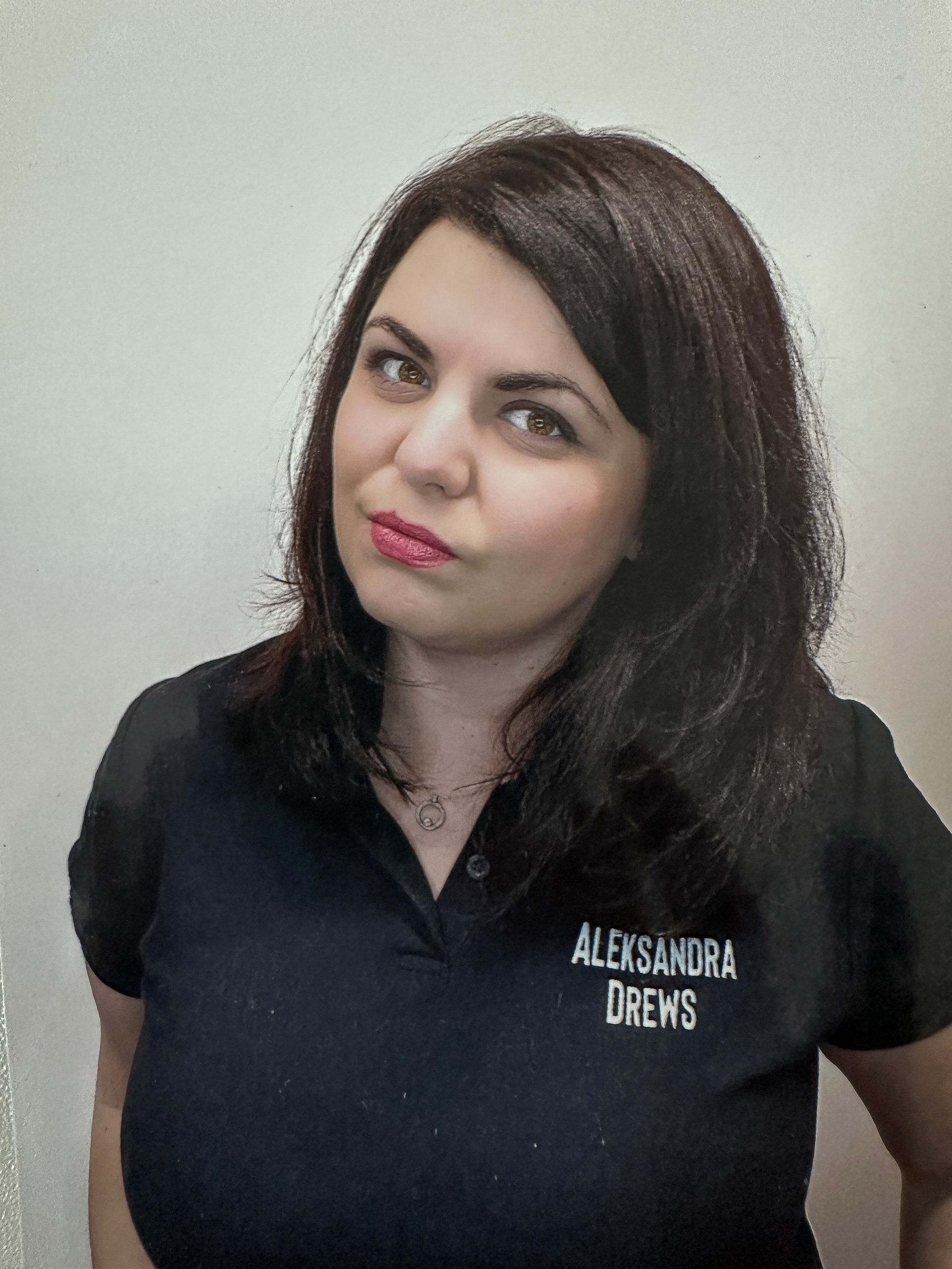 Frau mit dunklen Haaren in einem schwarzen Hemd; auf dem Logo steht „Aleksandra Drew’s“. Sie hat ein leichtes Lächeln im Gesicht.