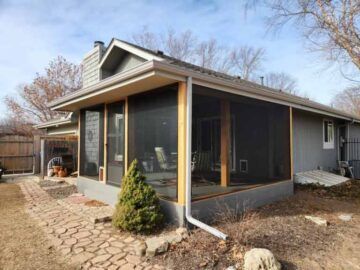 Screened porch attached to a gray house with a stone walkway and small evergreen in the yard
