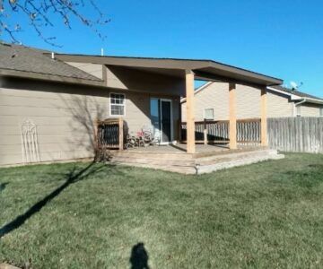 Backyard patio with beige house exterior, covered porch, and green lawn under blue sky