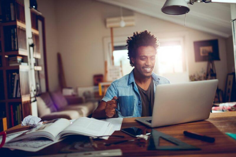Person smiling at a laptop in a cozy home office, with books and notebooks on a desk.