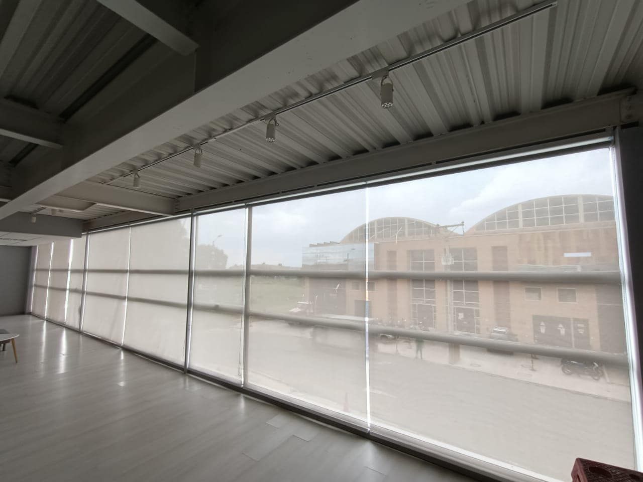 Vista interior: habitación luminosa con grandes ventanales, persianas y vistas a los edificios.
