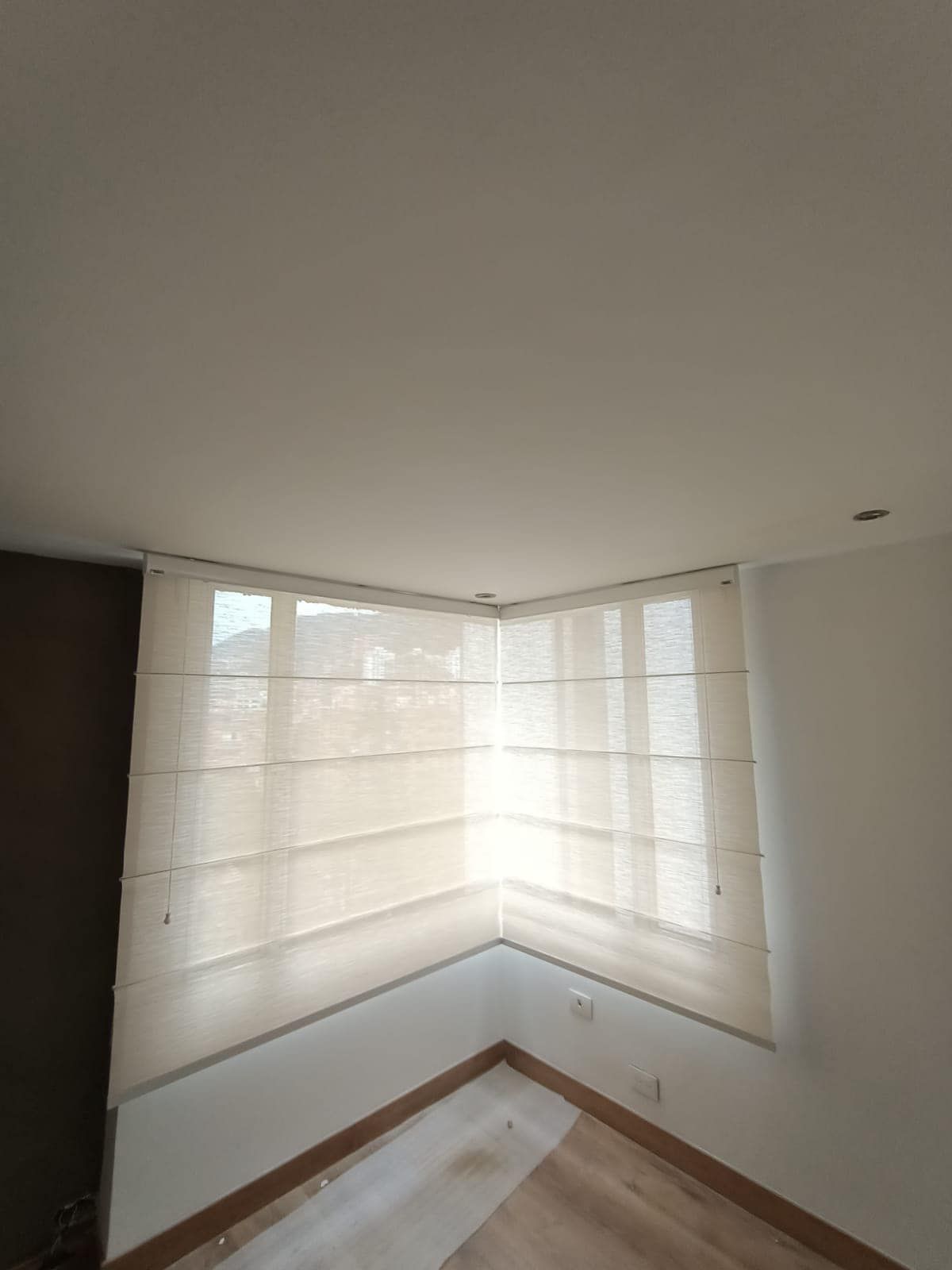 Ventana de esquina con cortinas romanas beige, paredes blancas y piso de madera.