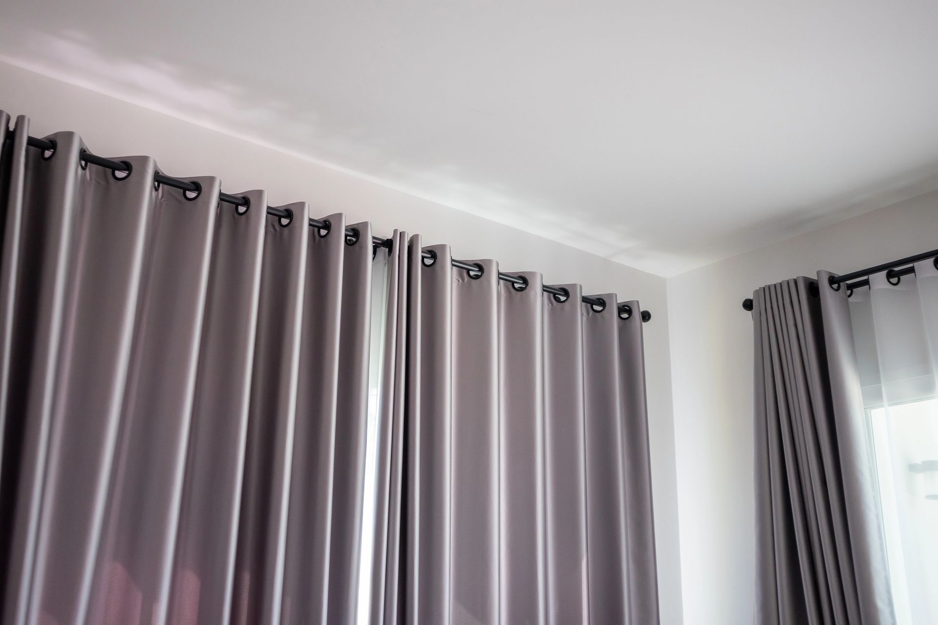 Cortinas grises con ojales colgadas de una barra de cortina contra una pared y techo blancos.