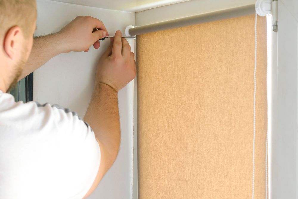 Persona instalando una persiana enrollable beige en un marco de ventana blanco, sosteniendo una herramienta.