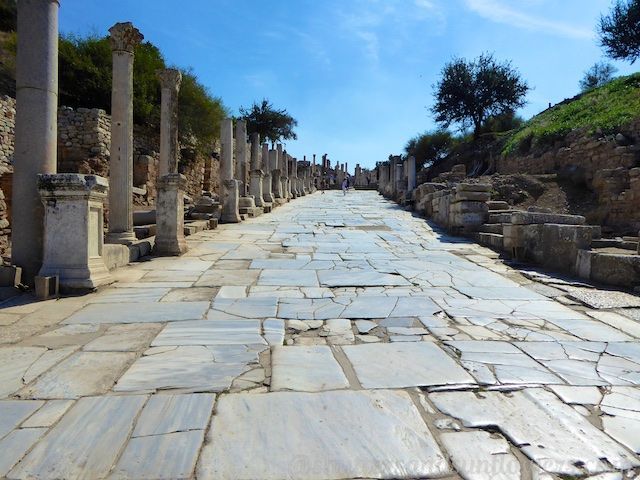 Stone road lined with columns, leading into distance under a blue sky, ruins of an ancient city.