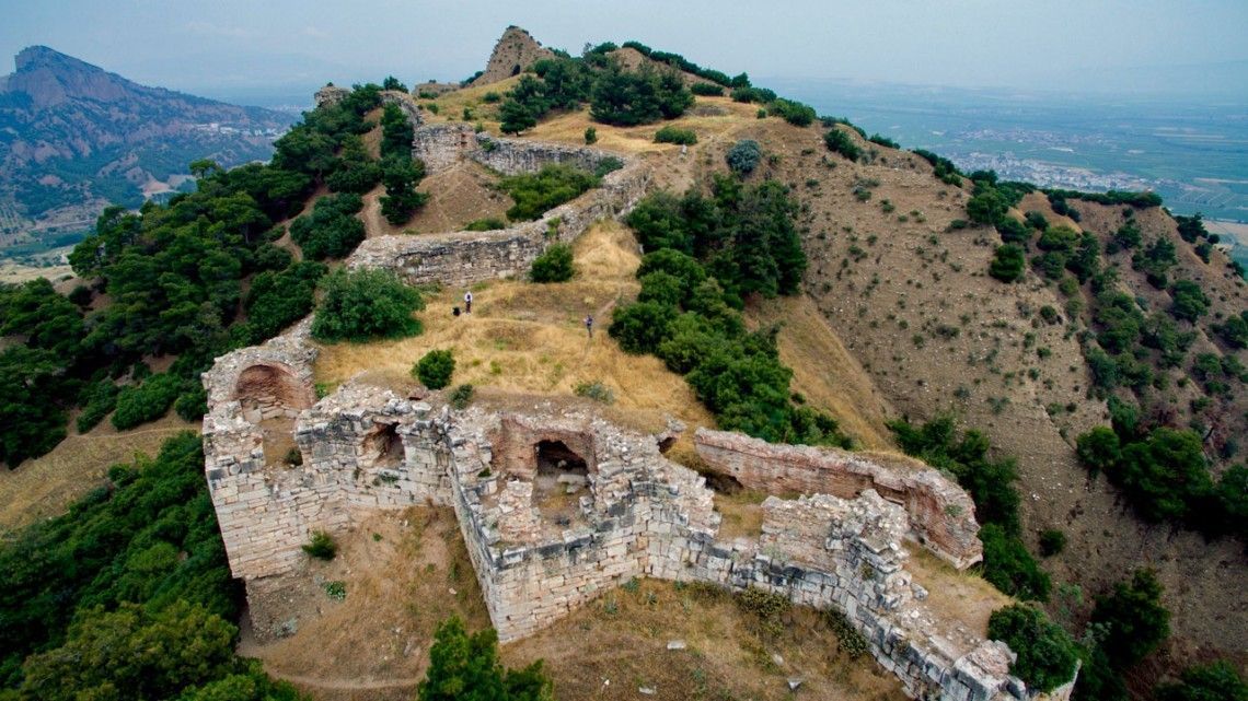 Ruin of a stone fortress atop a hilly landscape, with crumbling walls and surrounding greenery.