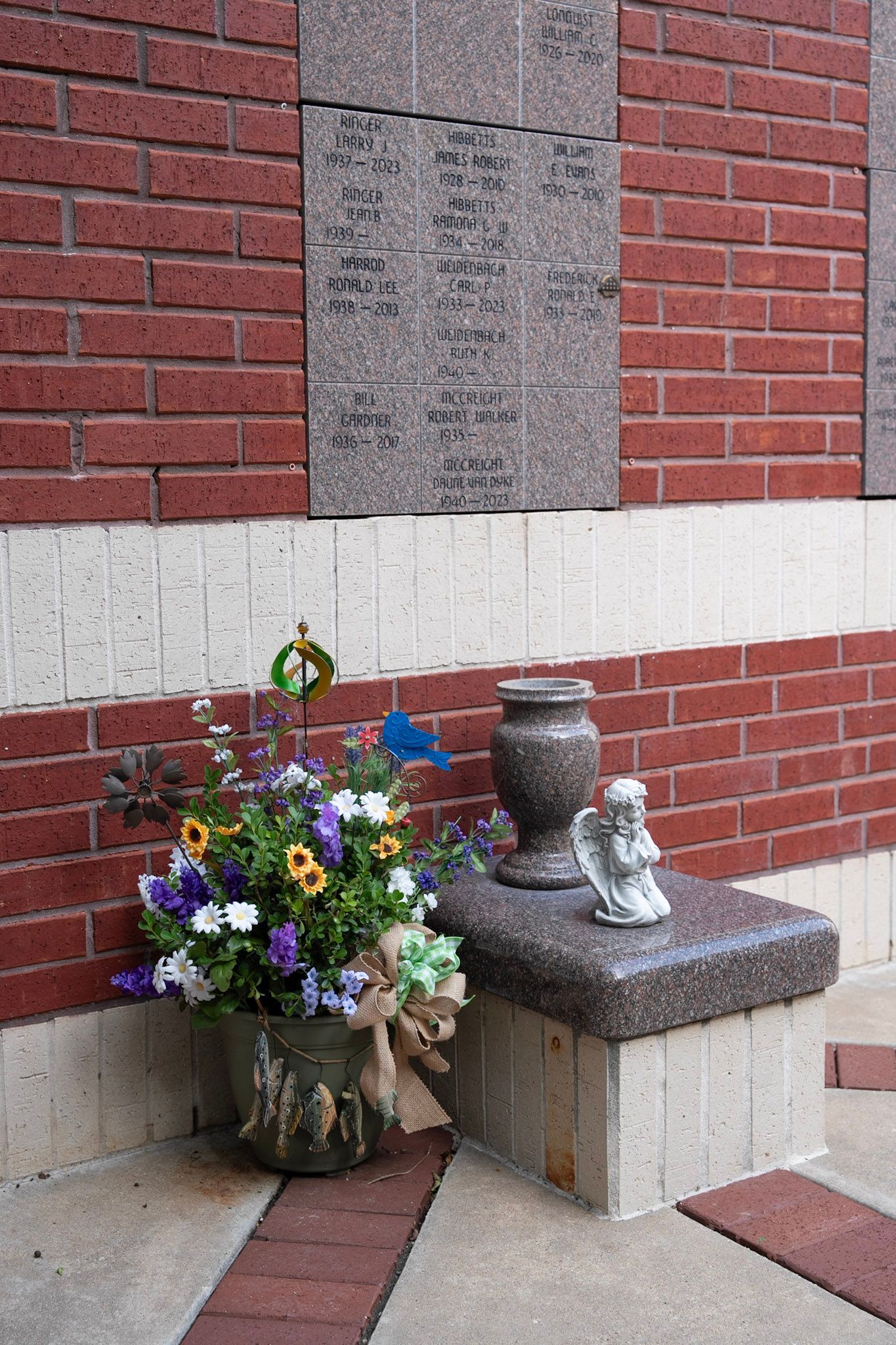 A brick wall with floral tributes, urn, and angel statue resting on a stone ledge at Christ Church Columbarium