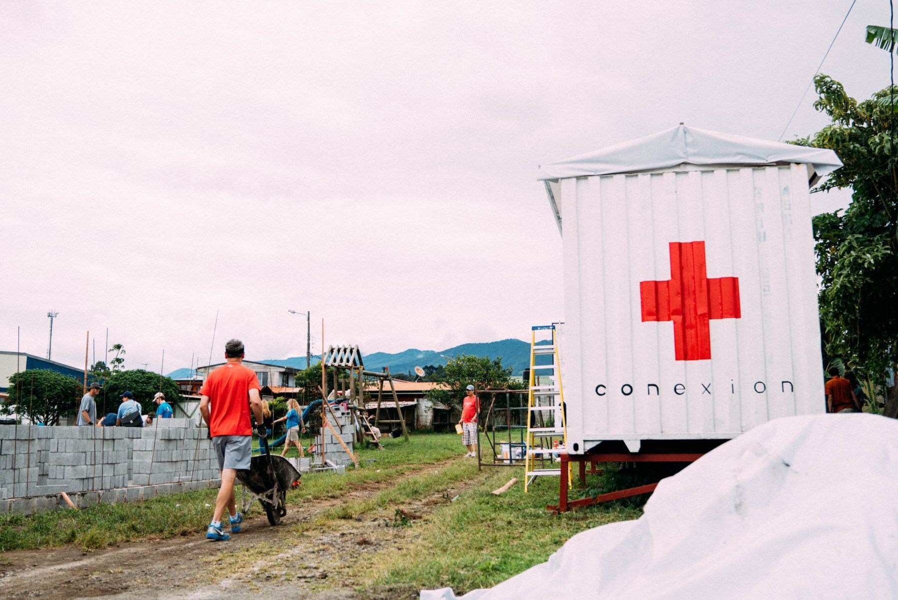 A man is walking down a dirt road in front of a white building with a red cross on it.