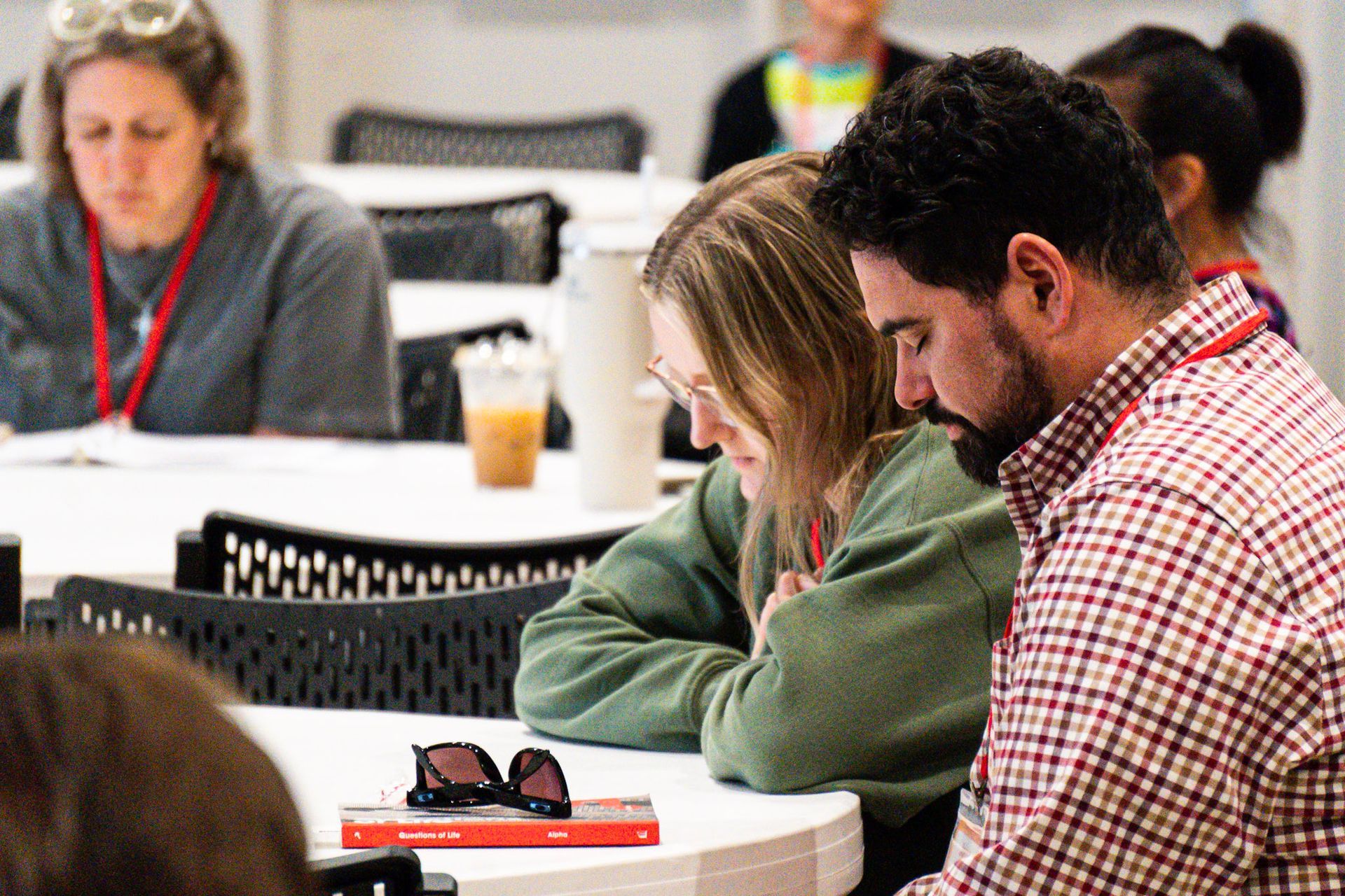 People at a table, looking down. One man in plaid shirt, one woman in green sweater, and another in the background.