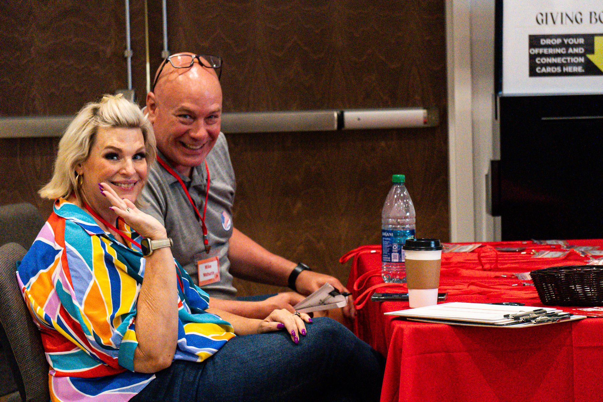 Woman in colorful shirt and man with glasses smile near a red table, water bottle and coffee cup.