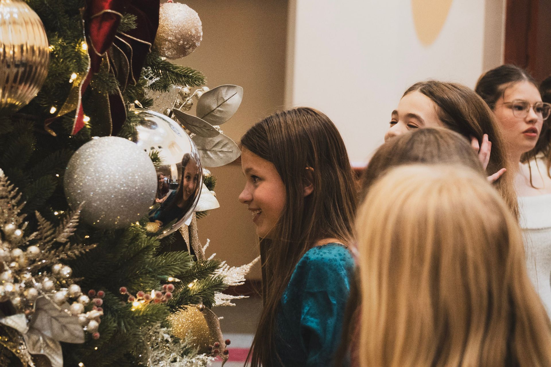 Girls admiring a decorated Christmas tree with ornaments; one smiles.