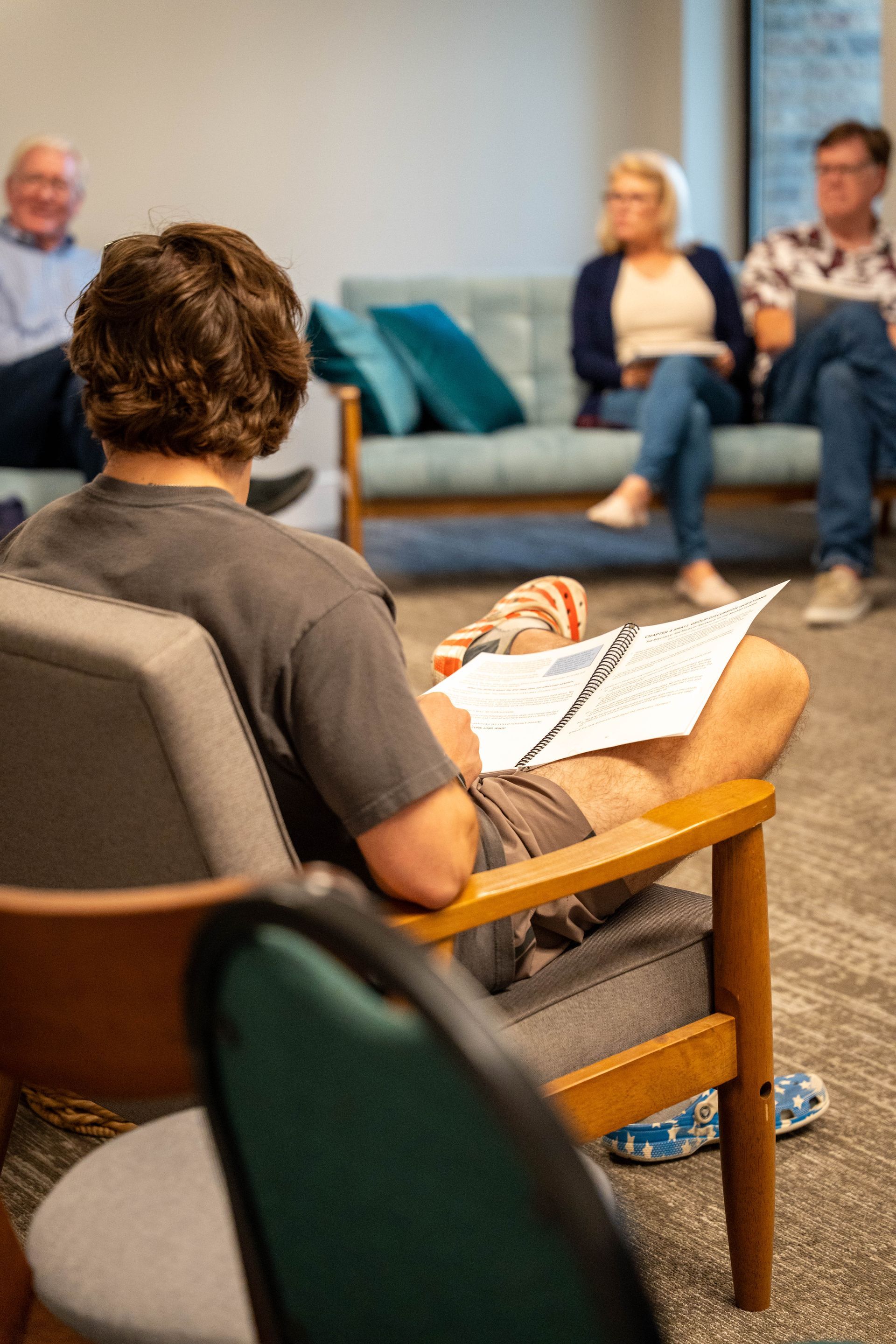 Man in gray shirt sits, looking at notes.  Other people on blue sofa, in a room.