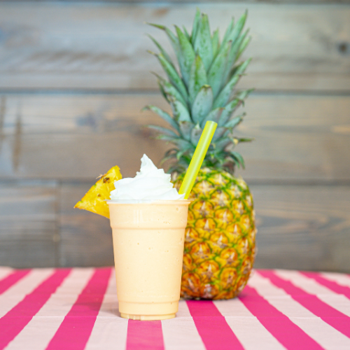 A pineapple smoothie with whipped cream and a straw is on a table next to a pineapple.