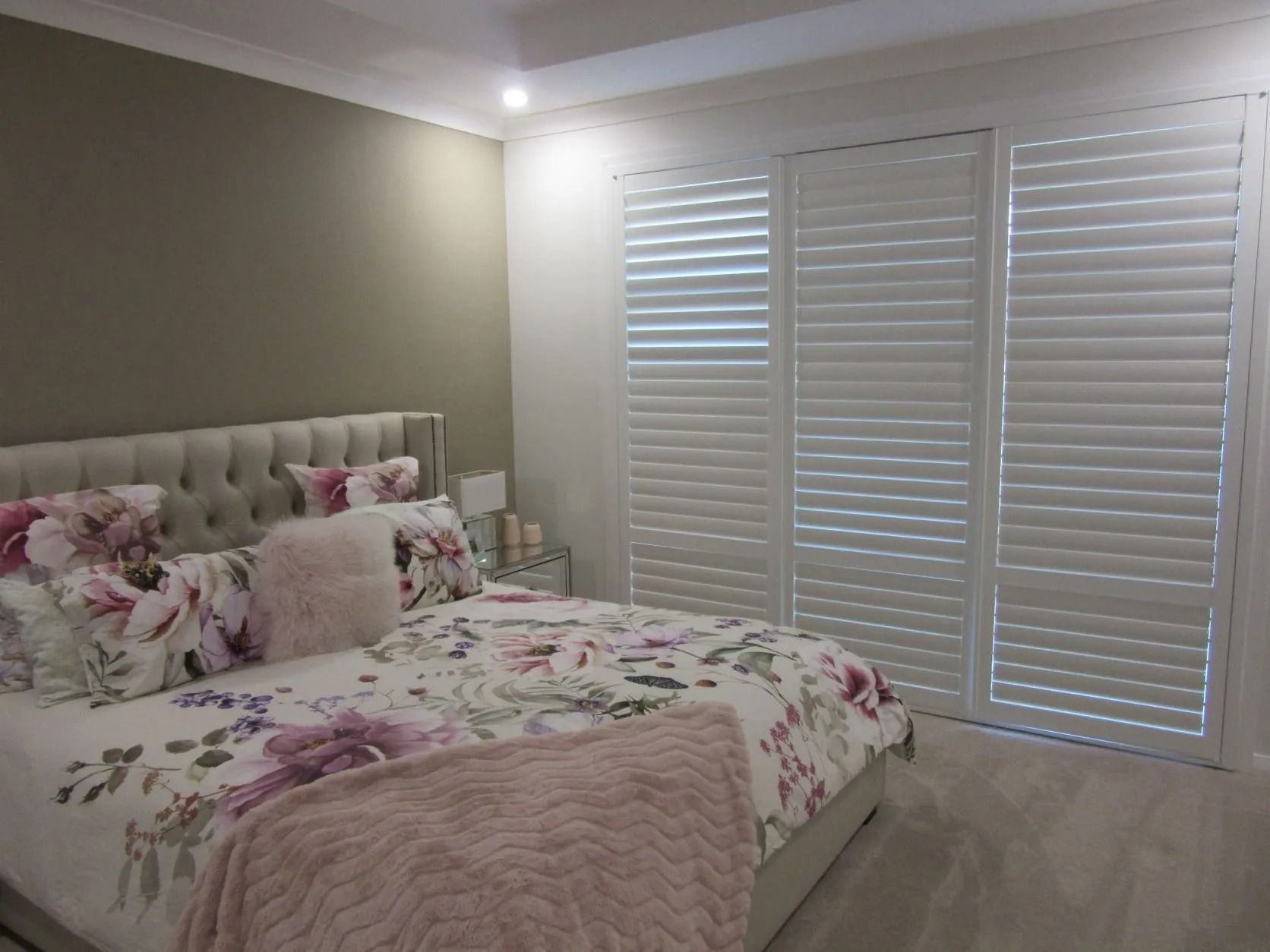 Bright bedroom with floral bedspread, tufted headboard, and white plantation shutters on the windows