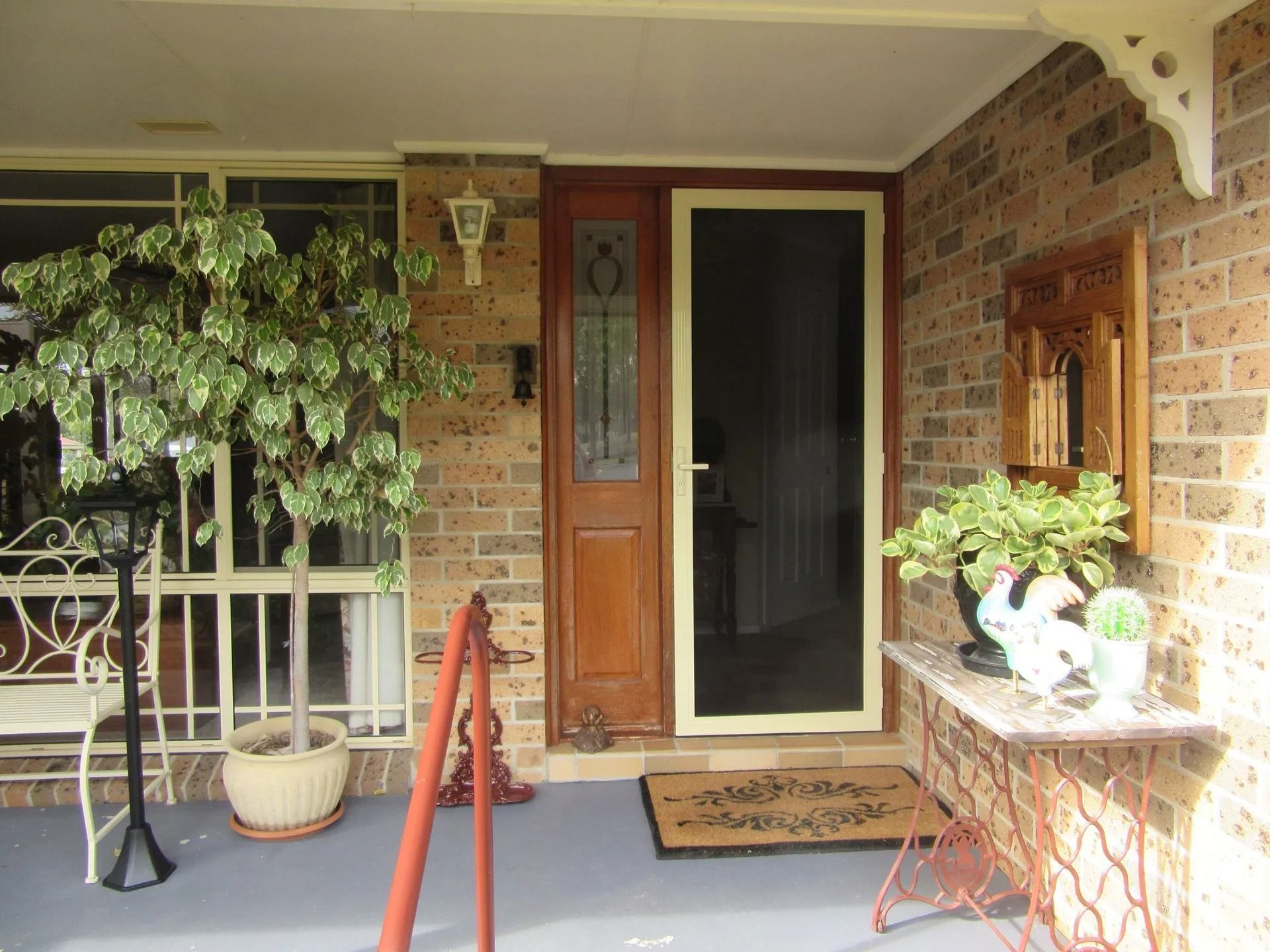 Brick front porch with wooden door, screen door, potted plants, and a small table with white decor pieces