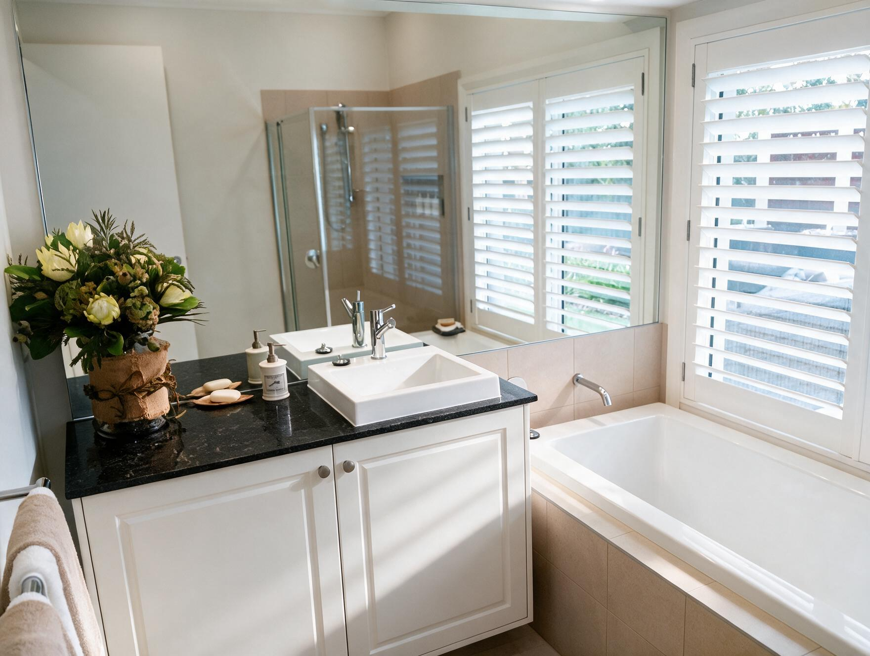 Bright bathroom with white vanity, black countertop, sink, bathtub, and plantation shutters on windows.