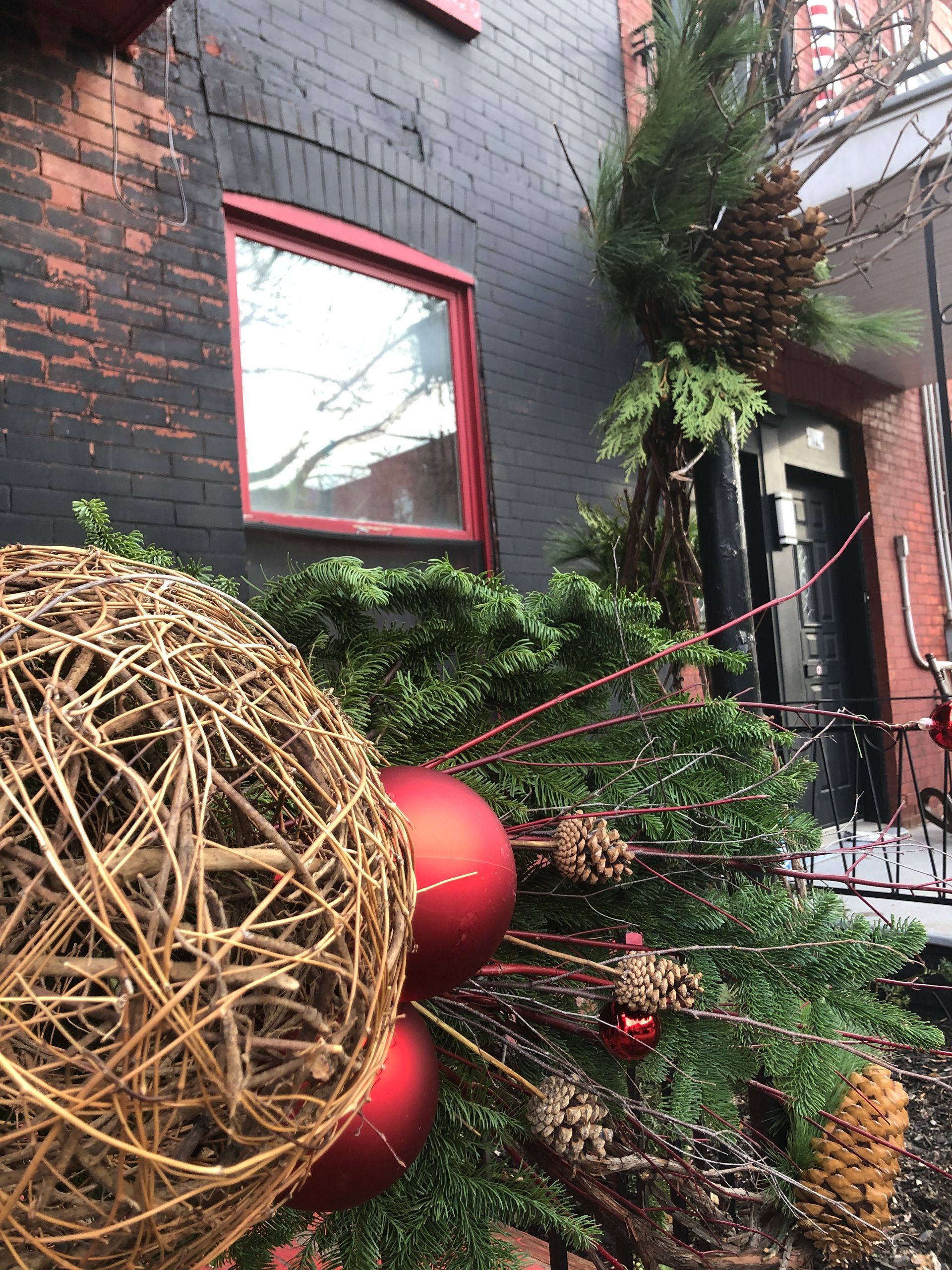 Un sapin de Noël avec des boules rouges et des pommes de pin devant un bâtiment en briques.