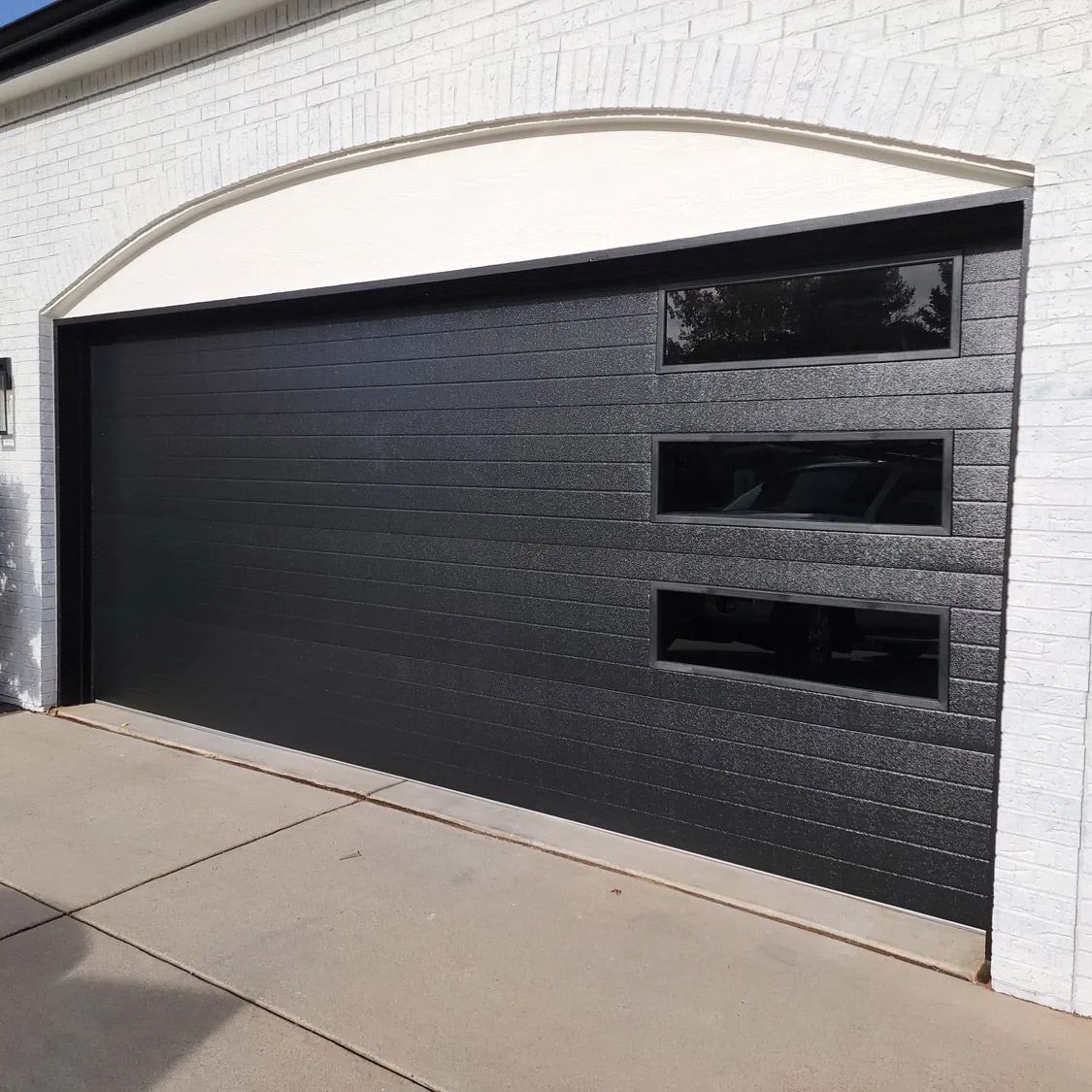 Black painted garage door with three narrow windows set in a white brick exterior wall