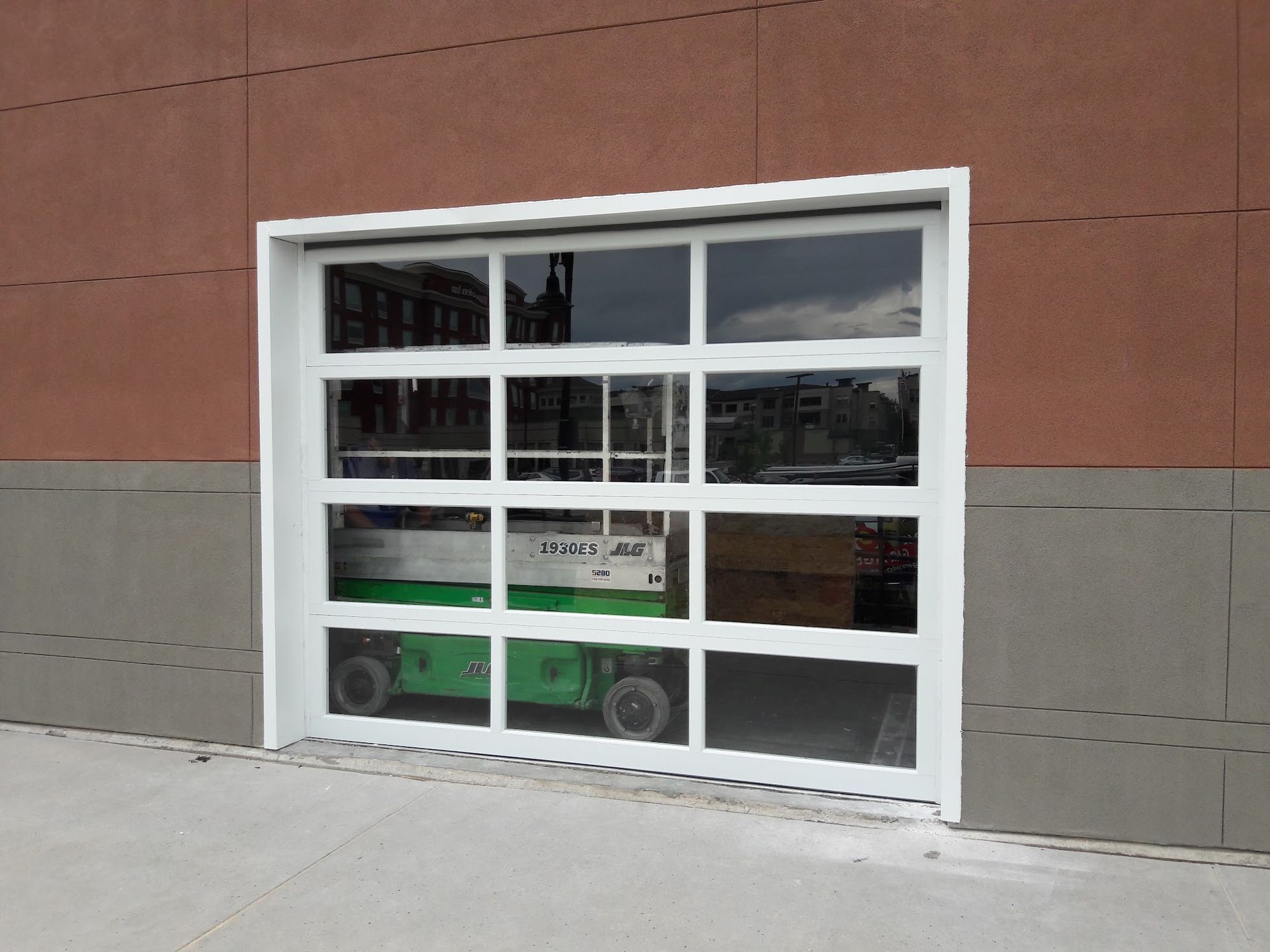 White-framed glass garage door revealing a green lift inside a building with red brick and grey trim.