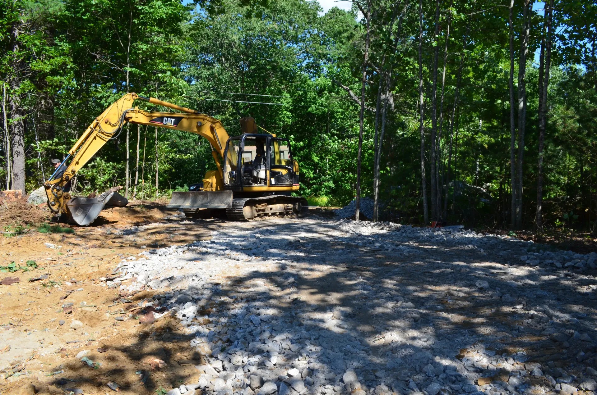 A yellow excavator is digging a gravel road in the woods.