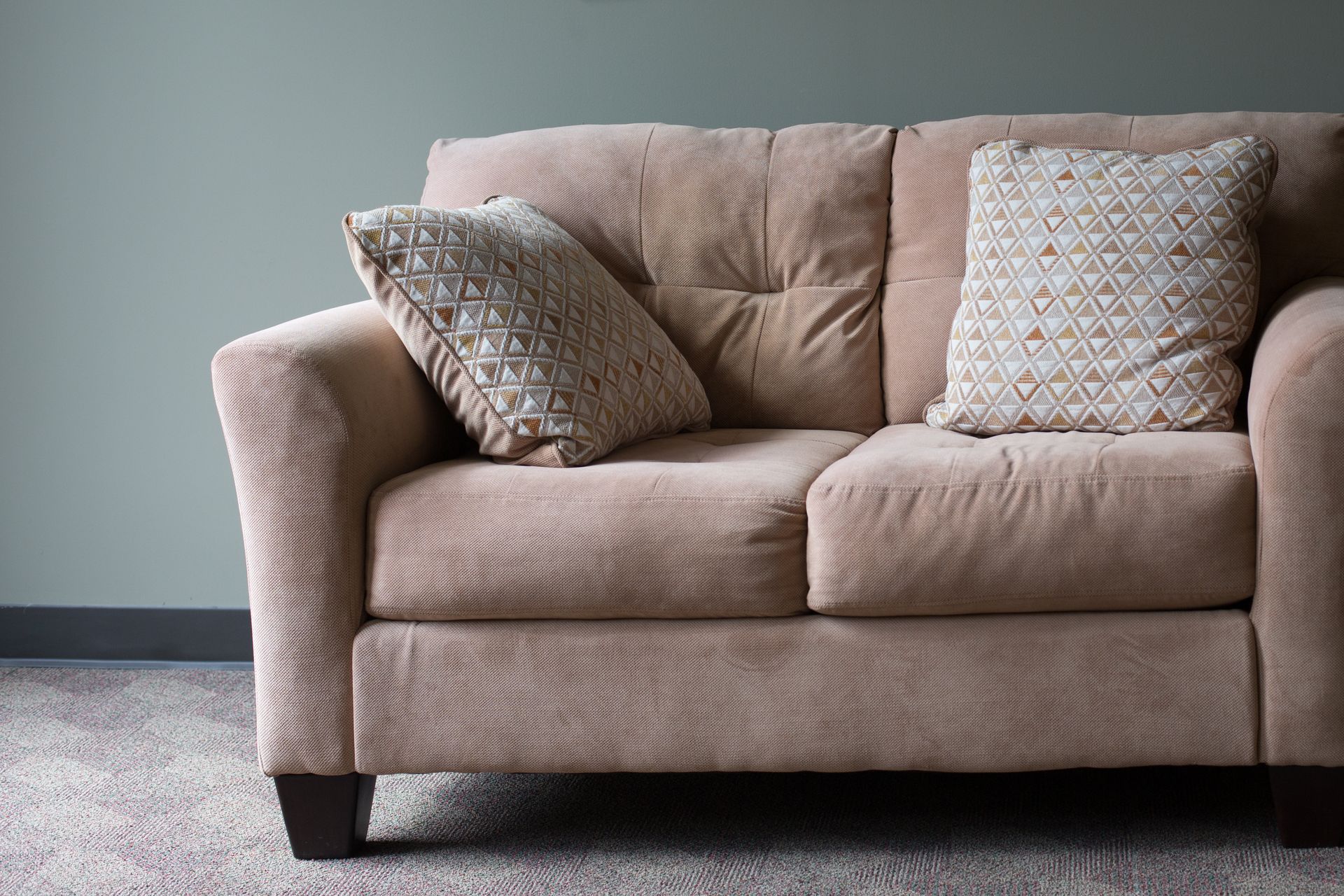 Beige loveseat with patterned pillows against a gray-green wall. Carpeted floor.