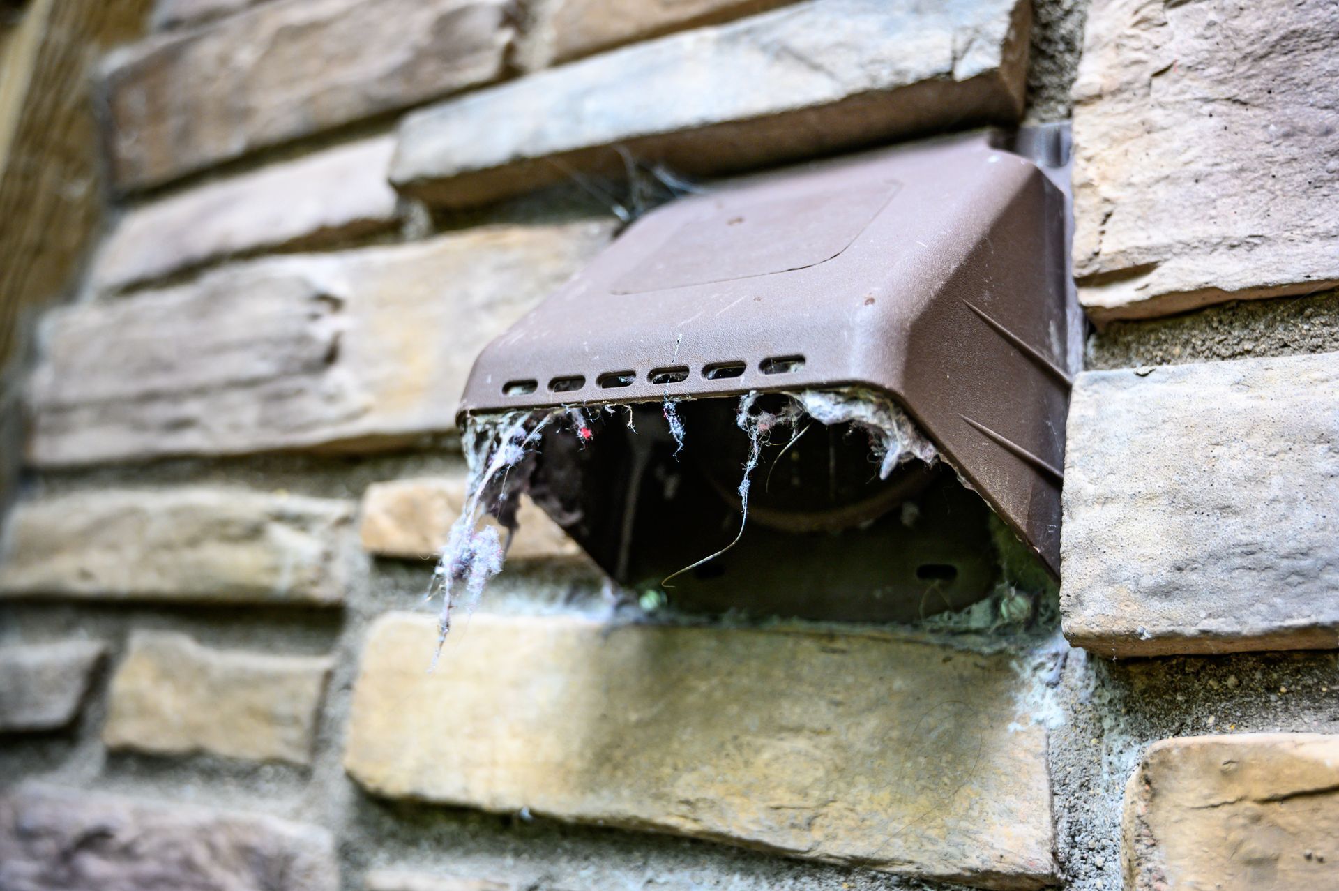 Brown dryer vent clogged with lint on a stone brick wall.